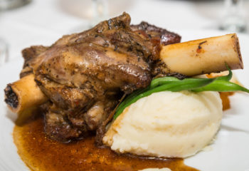 A veal or lamb shank on a plate with gravy, mashed potatoes, green beans and baby onions